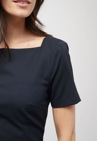 Marineblaues Kurzarmkleid mit quadratischem Ausschnitt. Hergestellt aus strukturiertem Stoff, ausgestattet mit einer diskreten Seitentasche und minimalistischen Designdetails.