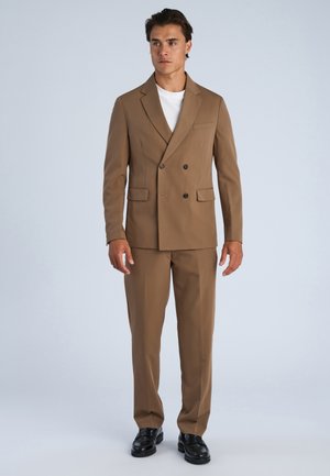 Mężczyzna ubrany w brązowy dwurzędowy garnitur z dopasowanymi spodniami i czarnymi eleganckimi butami, stojący na tle jednolitego jasnego tła.