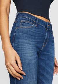Tmavě modré džíny z denimu s klasickým pětipocket designem, viditelným prošíváním a středním pasem. Tkanina vypadá hladce s mírnou texturou.