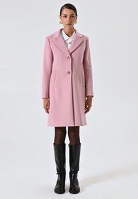 Donna in un cappotto rosa fino al ginocchio, blusa bianca, collant neri trasparenti e stivali neri fino al ginocchio, in piedi contro uno sfondo chiaro e semplice.