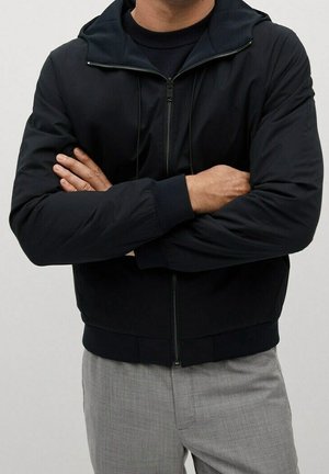 Homme aux bras croisés portant une veste noire zippée, un t-shirt noir en dessous et un pantalon gris clair, sur un fond clair uni.