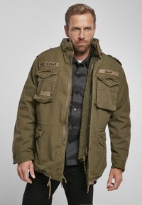 Olive green military-style jacket with multiple front pockets, a high collar, and a soft fleece lining. Features a drawstring hem and textured patches.