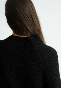 Mustan neulotun villapaidan, jossa on ribbikuvio, korkea kaula-aukko ja huomaamattomat vinonauhat selkäpuolella.