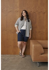 Chemise à rayures boutonnée bleu clair et blanc, portée sur un débardeur blanc. Short bleu marine avec cordon de serrage, associé à des tongs noires. Fond brun.