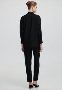 Maglietta nera a maniche lunghe con colletto, abbinata a pantaloni neri su misura. Tessuto liscio, vestibilità rilassata e design minimalista.