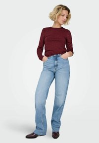 Kobieta o krótkich blond włosach, ubrana w bordową bluzkę z długim rękawem w kropki, jasnoniebieskie jeansy z wysokim stanem oraz ciemne buty, ręce w kieszeniach.