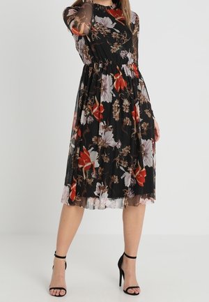 Woman wearing a black midi dress with sheer long sleeves and red, white, and brown floral patterns, paired with black ankle-strap high heels.