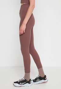 Barna, magas derekú leggings, rugalmas anyagból készült, feszes fazonú és minimális varrási részletekkel, fekete-fehér sportcipőkkel párosítva.