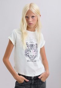 Tricou alb din bumbac cu mânecă scurtă, având o grafică cu fața unui tigru și textul "Născut să fiu liber" dedesubt, asortat cu șorturi negre din denim.