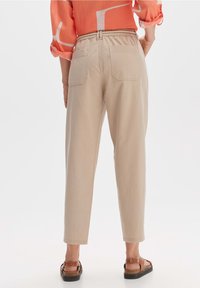 Beige taillierte Hose mit elastischem Bund, zwei Gesäßtaschen und einer glatten Stoffoberfläche. Kombiniert mit braunen Sandalen und einem korallenfarbenen Oberteil.
