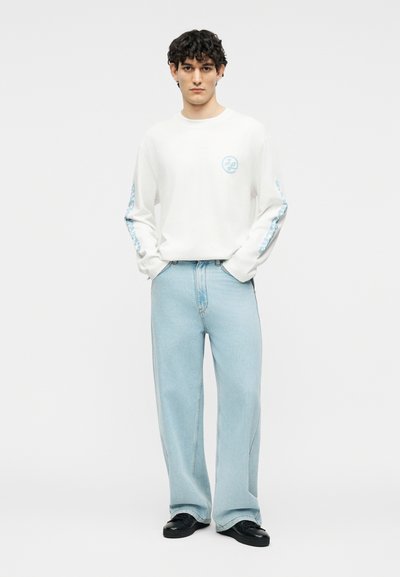 Άνδρας με σγουρά μαλλιά φοράει λευκό πουκάμισο με μακριά μανίκια και μπλε λογότυπο, ανοιχτό μπλε παντελόνι με φαρδιά μπατζάκια και μαύρα παπούτσια, στέκεται με τα χέρια στις τσέπες.