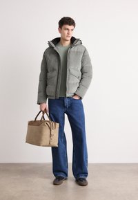Grå pufferjakke med ribbet tekstur, lynlås foran og to sidelommer. Paret med en lys grøn striktrøje, blå brede jeans og en tan taske.