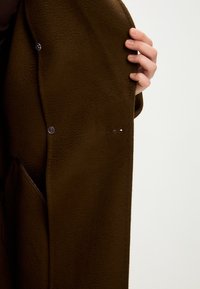 Brauner, strukturierter Stoffmantel mit einer Knopfverschlus und seitlichen Taschen. Der Ärmel zeigt ein dezentes Logo-Detail nahe dem Saum.