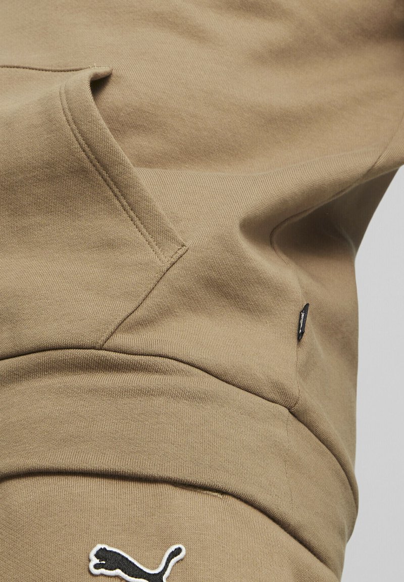 Felpa beige con tasca a marsupio, tessuto morbido e logo Puma bianco sul davanti. Presenta un piccolo etichetta nera con dettagli.