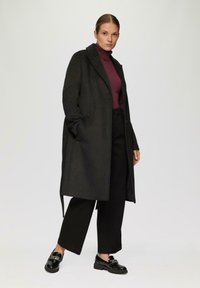 Zwarte lange jas met een gekarteld kraag, gecombineerd met een bordeauxrode coltrui, zwarte wijde broek en glanzende zwarte instapschoenen. Soepele textuur overal.