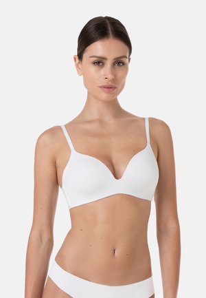 Donna con capelli scuri che indossa un reggiseno bianco e biancheria intima abbinata, in piedi davanti a uno sfondo chiaro e uniforme, rivolta in avanti.