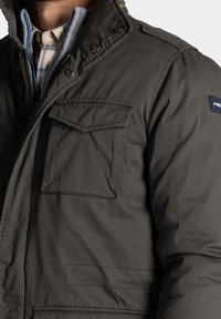 Donkergroene jas met een hoge kraag, drukknoopsluitingen en twee borstzakken. Beschikt over een gestructureerde stof en een gelaagde look met een geruit overhemd eronder.