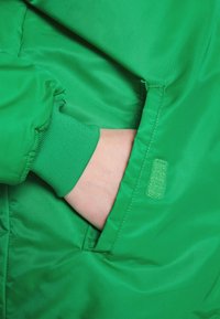Main portant une manchette en tricot vert insérée dans la poche latérale d'une veste vert vif avec des détails de coutures surpiquées.