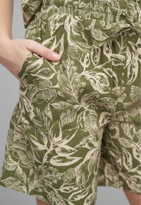 Shorts en tissu vert avec un imprimé crémeux de feuilles, taille élastique, poches latérales et coupe décontractée. Matériau naturel et texturé.