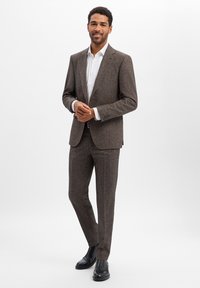 Brązowy garnitur z tweedu o dopasowanym kroju, z jednorzędową marynarką i spodniami z prostą nogawką. Połączony z białą koszulą i czarnymi butami.