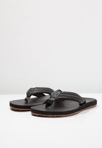 Svarte flip-flops med teksturerte skinnremmer, en flat gummisåle og brune detaljer. Har et diamantmønster på remmen.