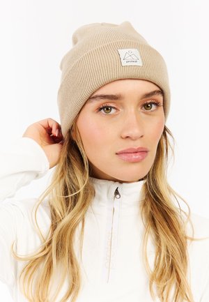 Young woman with long blonde hair wearing a beige knit beanie and white zip-up jacket, touching the back of the beanie.