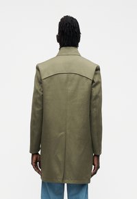 Palton verde măsliniu cu guler structurat, mâneci lungi și croială dreaptă, având o textură netedă și detalii minime de cusături.