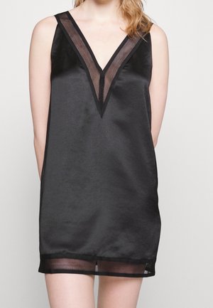 Czarna satynowa sukienka bez rękawów z siateczkowym dekoltem w kształcie litery V i siateczkowym dołem, noszona przez osobę o jasnej karnacji i blond włosach.