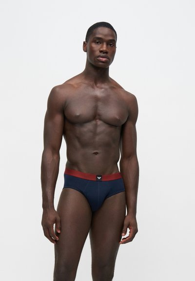 Homme musclé portant un slip bleu foncé avec une ceinture rouge, debout sur un fond blanc uni.