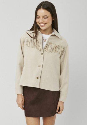 Veste beige avec un détail à franges sur le haut devant, dotée d'un large col et d'une fermeture à boutons. Texture douce, associée à une jupe marron.