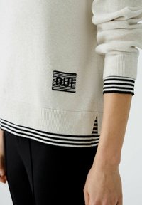 Helles beiges Sweatshirt mit schwarz-weißen gestreiften Bündchen. Verfügt über ein kleines aufgesticktes Patch mit dem Wort "OUT" und seitlichen Schlitzen am Saum.