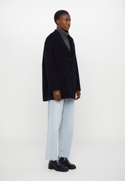 Manteau en mélange de laine bleu marine avec deux boutons, poches latérales et coupe décontractée, associé à un jean taille haute bleu clair à jambes larges et des bottes noires épaisses.