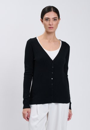 Frau mit zurückgebundenen dunklen Haaren, trägt eine schwarze knöpfbare Strickjacke über einem weißen Oberteil und weißen Hosen, stehend vor hellem Hintergrund.