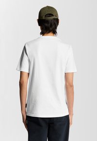 Hvid bomulds-t-shirt med korte ærmer og rund hals, set fra ryggen. Matcher mørke bukser og en olivengrøn cap.