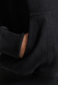 Hand in die Tasche eines schwarzen strukturierten Pullis mit gerippten Bündchen und Saum.