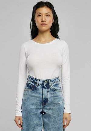 Weißes Langarmoberteil mit rundem Ausschnitt, kombiniert mit hoch taillierten, ausgewaschenen blauen Jeans. Der Schnitt ist schmal und das Material wirkt weich.