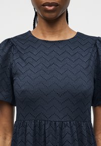 Γυναίκα που φοράει ένα μπλε σκούρο φόρεμα με κοντά μανίκια, σχέδιο ζιγκ-ζαγκ με τρύπες και με σούρα στη μέση.