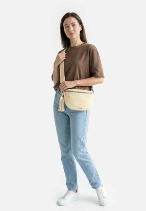 Femme portant un t-shirt marron, un jean bleu clair, des baskets blanches, avec un sac bandoulière beige orné de pompons et une sangle réglable.