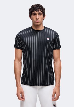 Uomo che indossa una camicia nera a maniche corte con strisce verticali bianche e pantaloni bianchi, in piedi di fronte a uno sfondo semplice.