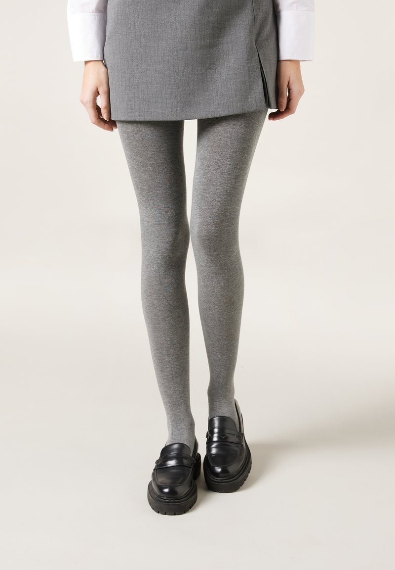 Grijze gebreide leggings met een gladde textuur, gecombineerd met zwarte loafers met een dikke zool. De outfit bevat een grijze rok.