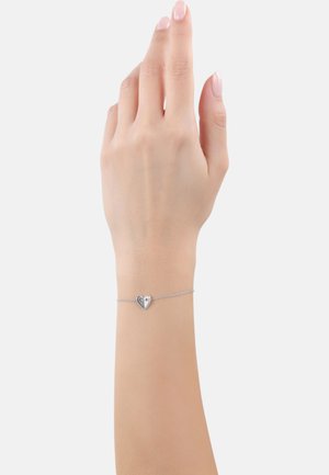 Braccialetto d'argento a forma di cuore su un polso, con una catena sottile e un ciondolo a forma di cuore dal design diviso, che riflette la luce.