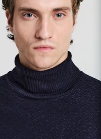 Marineblauer Rollkragenpullover mit strukturiertem Muster, geripptem Kragen und glattem Stoff. Nahaufnahme, die den Ausschnitt und die Gesichtszüge hervorhebt.