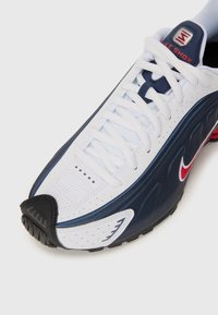 Nike Shox sneakers, med en hvit mesh-overdel med marineblå og røde detaljer, teksturerte paneler og en svart gummitåhette. Snøring.