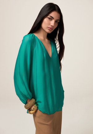 Blusa di seta turchese con scollatura a V, spalle arricciate e maniche lunghe. La modella indossa bracciali color oro sul polso.
