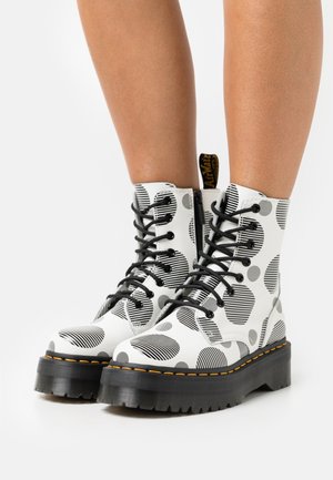 White ankle boots with black striped polka dot pattern, featuring black laces, a thick sole, and yellow stitching detail.