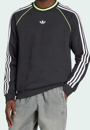 Persona que lleva una sudadera negra de Adidas con rayas blancas en las mangas y detalles en verde neón, combinada con pantalones grises de Adidas.