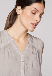 Hellgraues ärmelloses Blusenshirt mit V-Ausschnitt, feinen Falten und fünf Knopfverschluss. Weicher Stoff mit glatter Textur und zarter Nahtführung.