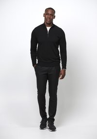 Schwarzer Pullover mit Reißverschluss am Hals und langen Ärmeln, kombiniert mit eng anliegenden schwarzen Hosen und schwarzen Sportschuhen. Schlichte Stoffqualität, keine sichtbaren Muster.