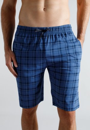 Mann trägt kniehohe Shorts in blau-schwarz kariert mit elastischem Bund und Kordelzug, steht mit einer Hand in der Tasche.