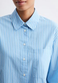 Camicia button-up azzurro chiaro con righe verticali bianche, colletto classico e tasca sul petto con una finitura in cotone testurizzato.